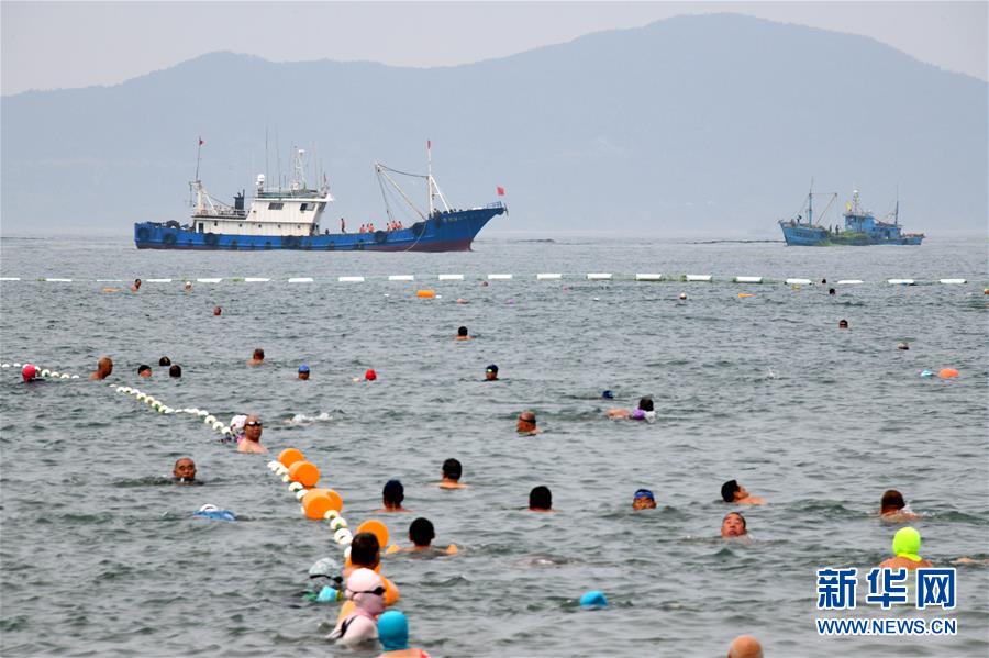 7月17日，游客在青岛第一海水浴场游泳，远处是打捞浒苔的渔船。